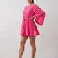 Woman wearing the Pleated Ruffle Detail Mini Dress featuring an all-over pleated design, flowing bell sleeves, a round neckline, elasticated waist with a self-tie sash, and a feminine ruffle hem, perfect for weddings, garden parties, race days, and elegant occasions.