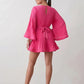 Woman wearing the Pleated Ruffle Detail Mini Dress featuring an all-over pleated design, flowing bell sleeves, a round neckline, elasticated waist with a self-tie sash, and a feminine ruffle hem, perfect for weddings, garden parties, race days, and elegant occasions.