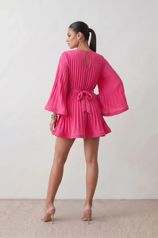 Woman wearing the Pleated Ruffle Detail Mini Dress featuring an all-over pleated design, flowing bell sleeves, a round neckline, elasticated waist with a self-tie sash, and a feminine ruffle hem, perfect for weddings, garden parties, race days, and elegant occasions.