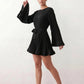 Woman wearing the Pleated Ruffle Detail Mini Dress featuring an all-over pleated design, flowing bell sleeves, a round neckline, elasticated waist with a self-tie sash, and a feminine ruffle hem, perfect for weddings, garden parties, race days, and elegant occasions.