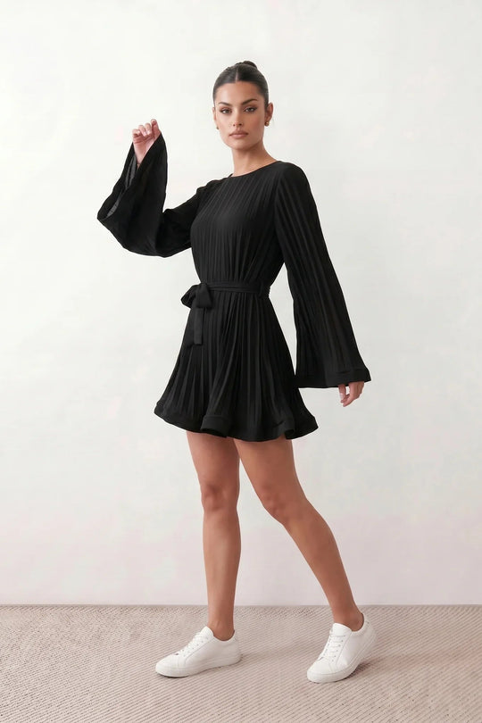 Woman wearing the Pleated Ruffle Detail Mini Dress featuring an all-over pleated design, flowing bell sleeves, a round neckline, elasticated waist with a self-tie sash, and a feminine ruffle hem, perfect for weddings, garden parties, race days, and elegant occasions.