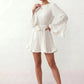 Woman wearing the Pleated Ruffle Detail Mini Dress featuring an all-over pleated design, flowing bell sleeves, a round neckline, elasticated waist with a self-tie sash, and a feminine ruffle hem, perfect for weddings, garden parties, race days, and elegant occasions.