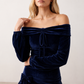 Woman wearing the Silviana Glossy Bardot Mini Dress featuring a glossy finish, off-shoulder Bardot neckline with soft shirring, centre front bow detail, long sleeves and a flattering dropped waist silhouette, styled for parties and evening occasions.