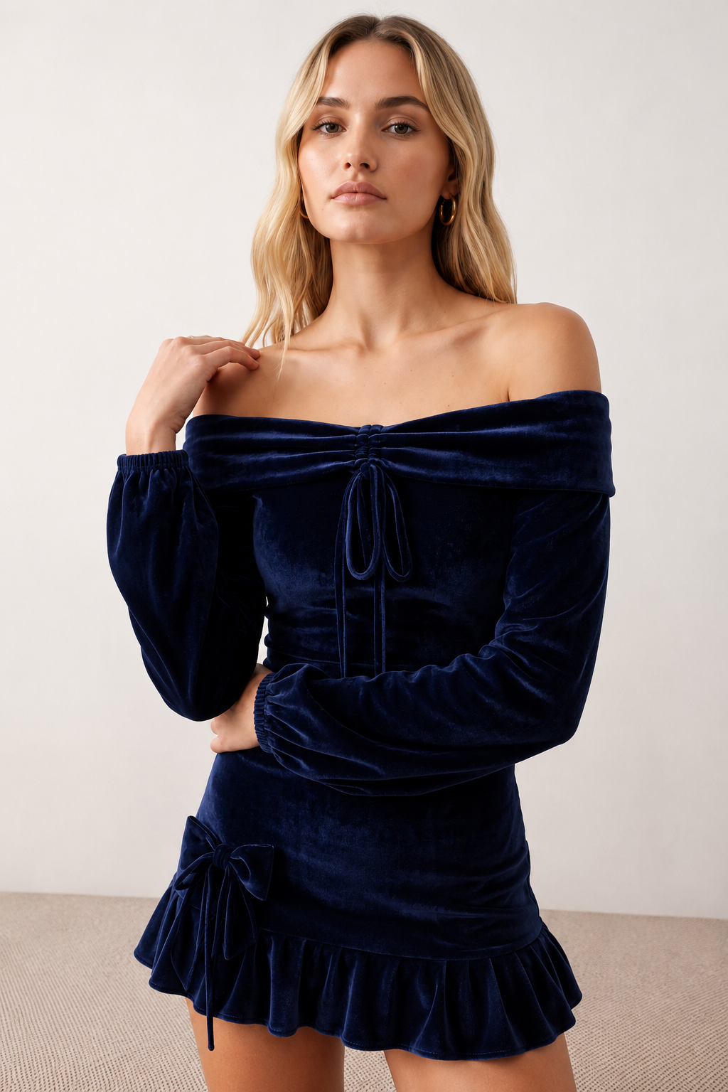 Woman wearing the Silviana Glossy Bardot Mini Dress featuring a glossy finish, off-shoulder Bardot neckline with soft shirring, centre front bow detail, long sleeves and a flattering dropped waist silhouette, styled for parties and evening occasions.