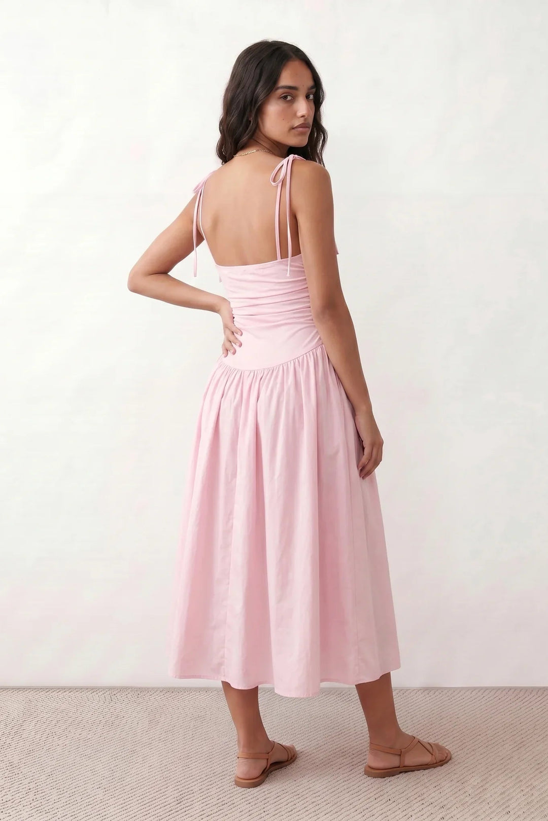 Woman wearing a spaghetti strap low back midi dress with a square neckline, ruched ruffle bodice, adjustable tie shoulder straps, elasticated smocked back, and flowing A-line skirt, styled as an elegant wedding guest and summer occasion dress.