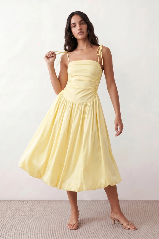 Woman wearing a spaghetti strap low back midi dress with a square neckline, ruched ruffle bodice, adjustable tie shoulder straps, elasticated smocked back, and flowing A-line skirt, styled as an elegant wedding guest and summer occasion dress.