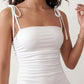 Woman wearing a spaghetti strap low back midi dress with a square neckline, ruched ruffle bodice, adjustable tie shoulder straps, elasticated smocked back, and flowing A-line skirt, styled as an elegant wedding guest and summer occasion dress.