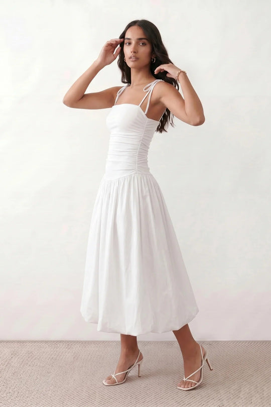 Woman wearing a spaghetti strap low back midi dress with a square neckline, ruched ruffle bodice, adjustable tie shoulder straps, elasticated smocked back, and flowing A-line skirt, styled as an elegant wedding guest and summer occasion dress.