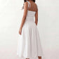 Woman wearing a spaghetti strap low back midi dress with a square neckline, ruched ruffle bodice, adjustable tie shoulder straps, elasticated smocked back, and flowing A-line skirt, styled as an elegant wedding guest and summer occasion dress.