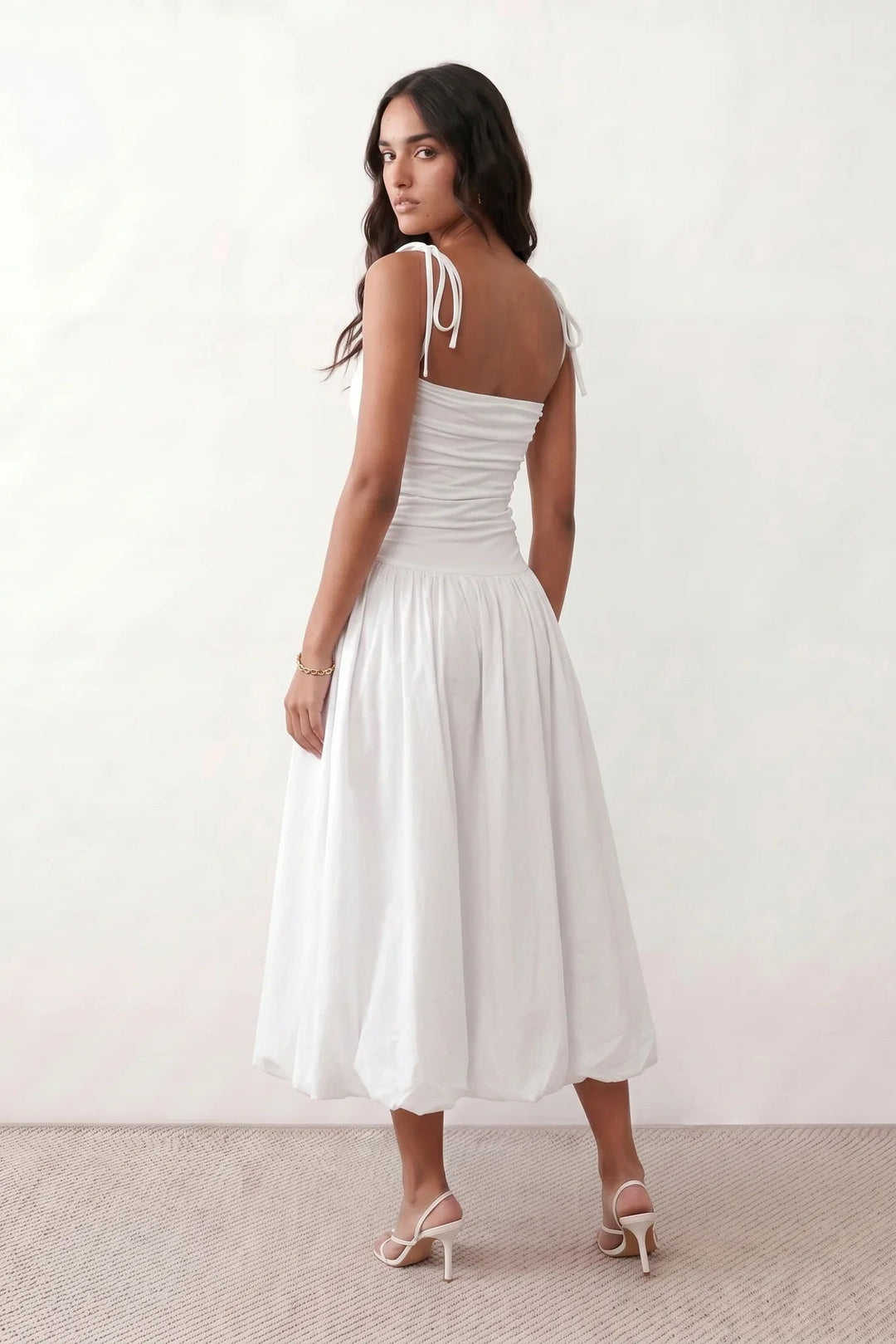 Woman wearing a spaghetti strap low back midi dress with a square neckline, ruched ruffle bodice, adjustable tie shoulder straps, elasticated smocked back, and flowing A-line skirt, styled as an elegant wedding guest and summer occasion dress.