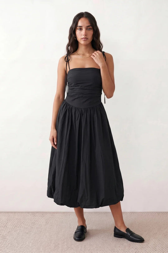 Woman wearing a spaghetti strap low back midi dress with a square neckline, ruched ruffle bodice, adjustable tie shoulder straps, elasticated smocked back, and flowing A-line skirt, styled as an elegant wedding guest and summer occasion dress.