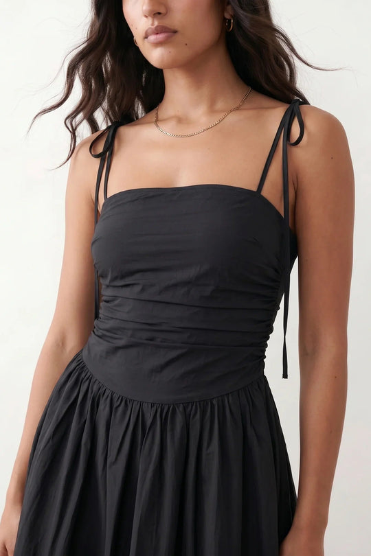 Woman wearing a spaghetti strap low back midi dress with a square neckline, ruched ruffle bodice, adjustable tie shoulder straps, elasticated smocked back, and flowing A-line skirt, styled as an elegant wedding guest and summer occasion dress.