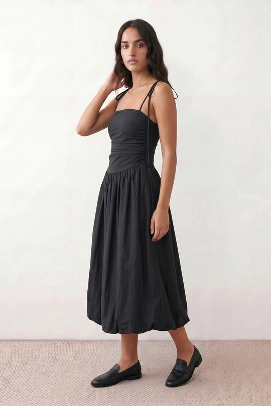 Woman wearing a spaghetti strap low back midi dress with a square neckline, ruched ruffle bodice, adjustable tie shoulder straps, elasticated smocked back, and flowing A-line skirt, styled as an elegant wedding guest and summer occasion dress.