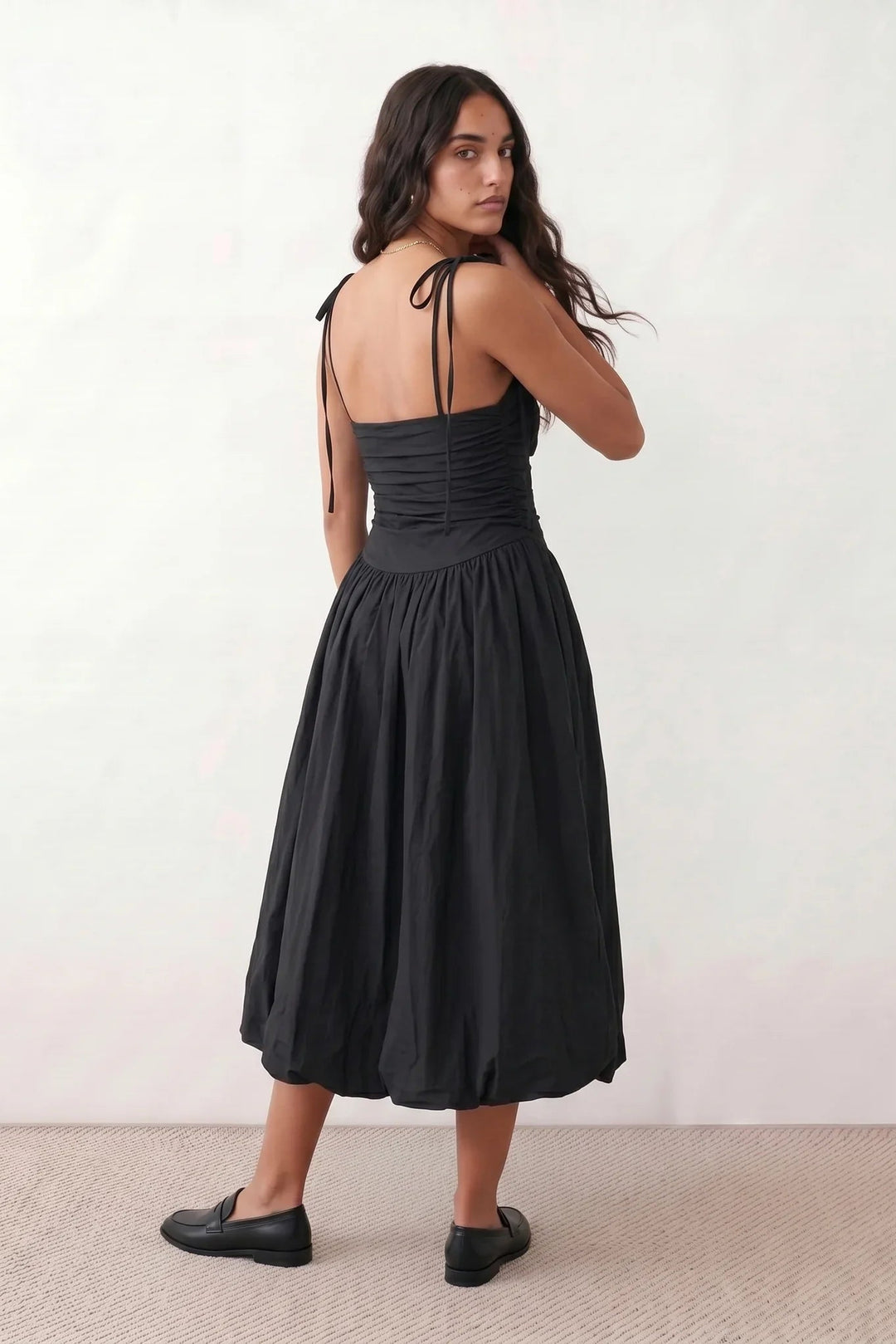 Woman wearing a spaghetti strap low back midi dress with a square neckline, ruched ruffle bodice, adjustable tie shoulder straps, elasticated smocked back, and flowing A-line skirt, styled as an elegant wedding guest and summer occasion dress.