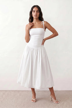Woman wearing a spaghetti strap low back midi dress with a square neckline, ruched ruffle bodice, adjustable tie shoulder straps, elasticated smocked back, and flowing A-line skirt, styled as an elegant wedding guest and summer occasion dress.