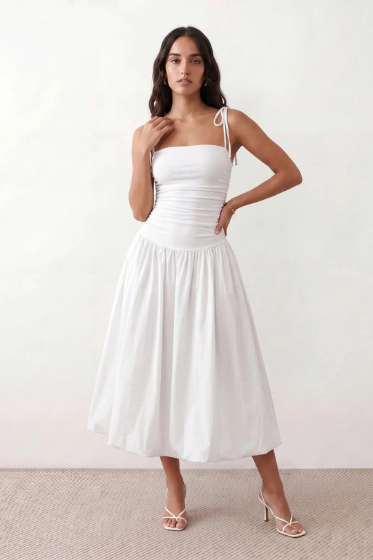 Woman wearing a spaghetti strap low back midi dress with a square neckline, ruched ruffle bodice, adjustable tie shoulder straps, elasticated smocked back, and flowing A-line skirt, styled as an elegant wedding guest and summer occasion dress.