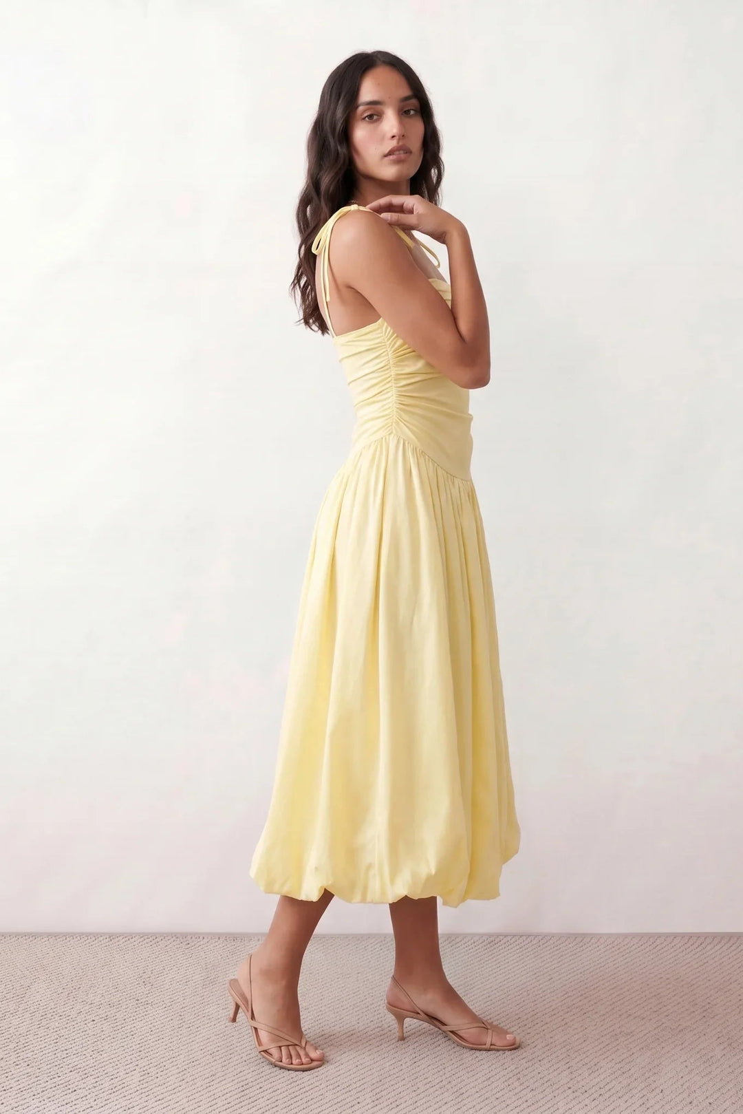 Woman wearing a spaghetti strap low back midi dress with a square neckline, ruched ruffle bodice, adjustable tie shoulder straps, elasticated smocked back, and flowing A-line skirt, styled as an elegant wedding guest and summer occasion dress.