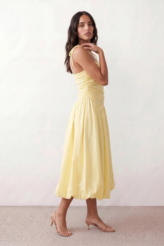 Woman wearing a spaghetti strap low back midi dress with a square neckline, ruched ruffle bodice, adjustable tie shoulder straps, elasticated smocked back, and flowing A-line skirt, styled as an elegant wedding guest and summer occasion dress.
