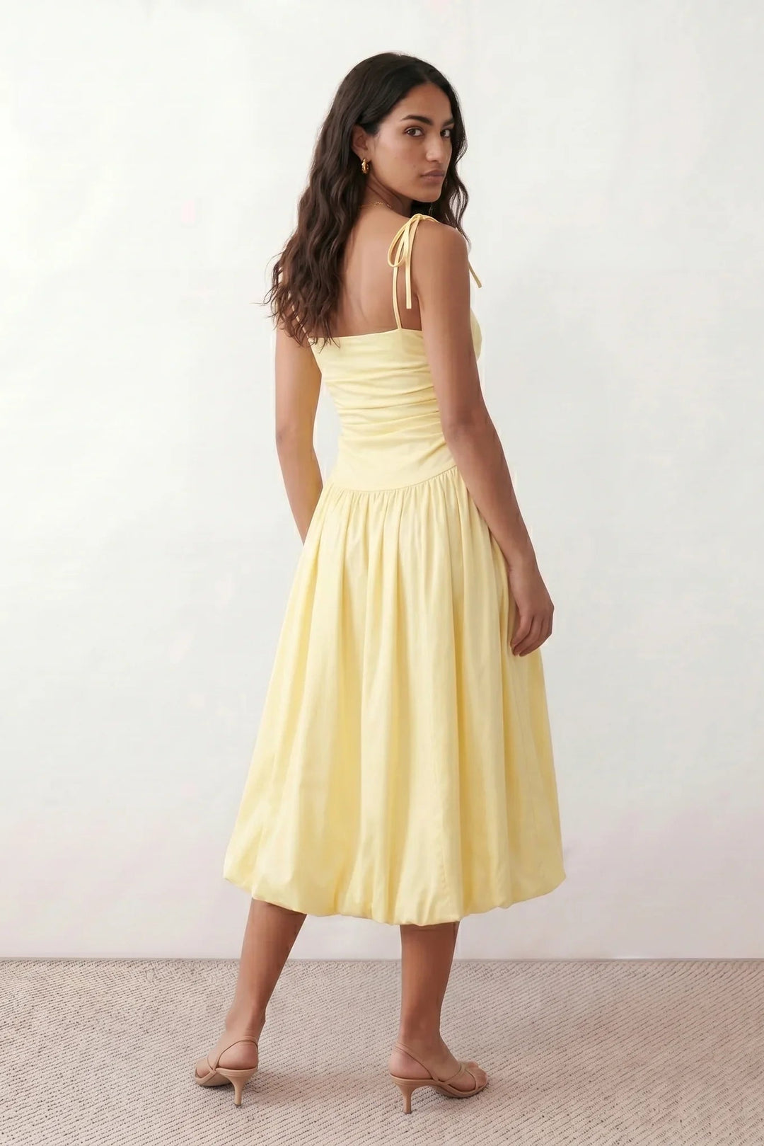 Woman wearing a spaghetti strap low back midi dress with a square neckline, ruched ruffle bodice, adjustable tie shoulder straps, elasticated smocked back, and flowing A-line skirt, styled as an elegant wedding guest and summer occasion dress.