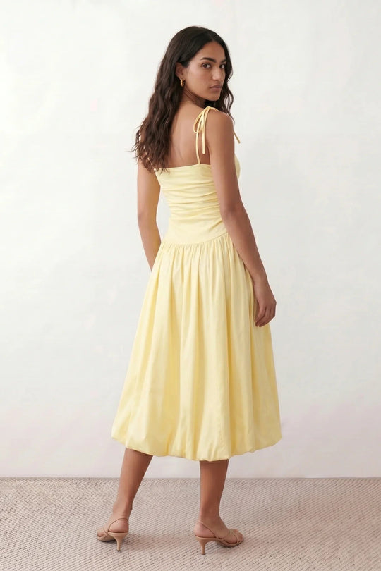 Woman wearing a spaghetti strap low back midi dress with a square neckline, ruched ruffle bodice, adjustable tie shoulder straps, elasticated smocked back, and flowing A-line skirt, styled as an elegant wedding guest and summer occasion dress.