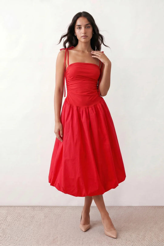 Woman wearing a spaghetti strap low back midi dress with a square neckline, ruched ruffle bodice, adjustable tie shoulder straps, elasticated smocked back, and flowing A-line skirt, styled as an elegant wedding guest and summer occasion dress.