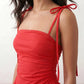 Woman wearing a spaghetti strap low back midi dress with a square neckline, ruched ruffle bodice, adjustable tie shoulder straps, elasticated smocked back, and flowing A-line skirt, styled as an elegant wedding guest and summer occasion dress.