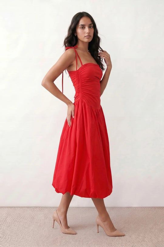 Woman wearing a spaghetti strap low back midi dress with a square neckline, ruched ruffle bodice, adjustable tie shoulder straps, elasticated smocked back, and flowing A-line skirt, styled as an elegant wedding guest and summer occasion dress.
