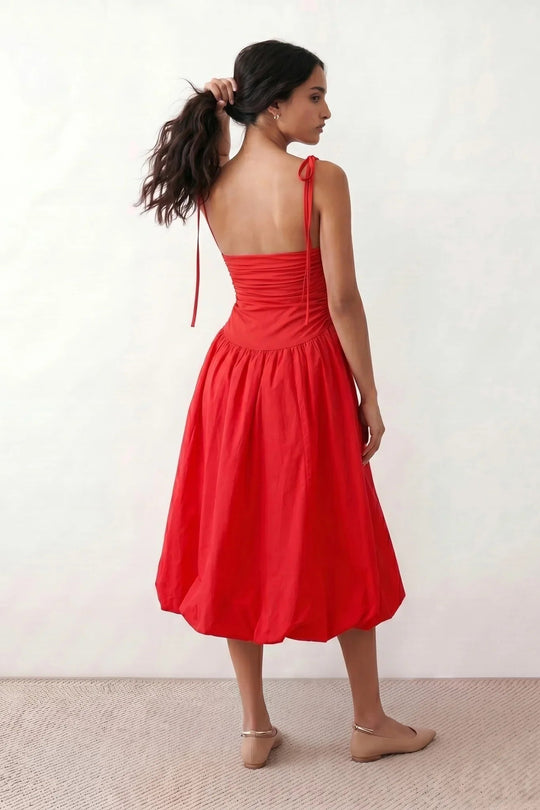 Woman wearing a spaghetti strap low back midi dress with a square neckline, ruched ruffle bodice, adjustable tie shoulder straps, elasticated smocked back, and flowing A-line skirt, styled as an elegant wedding guest and summer occasion dress.