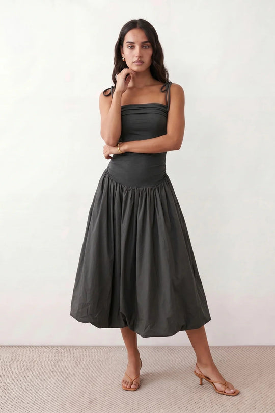 Woman wearing a spaghetti strap low back midi dress with a square neckline, ruched ruffle bodice, adjustable tie shoulder straps, elasticated smocked back, and flowing A-line skirt, styled as an elegant wedding guest and summer occasion dress.