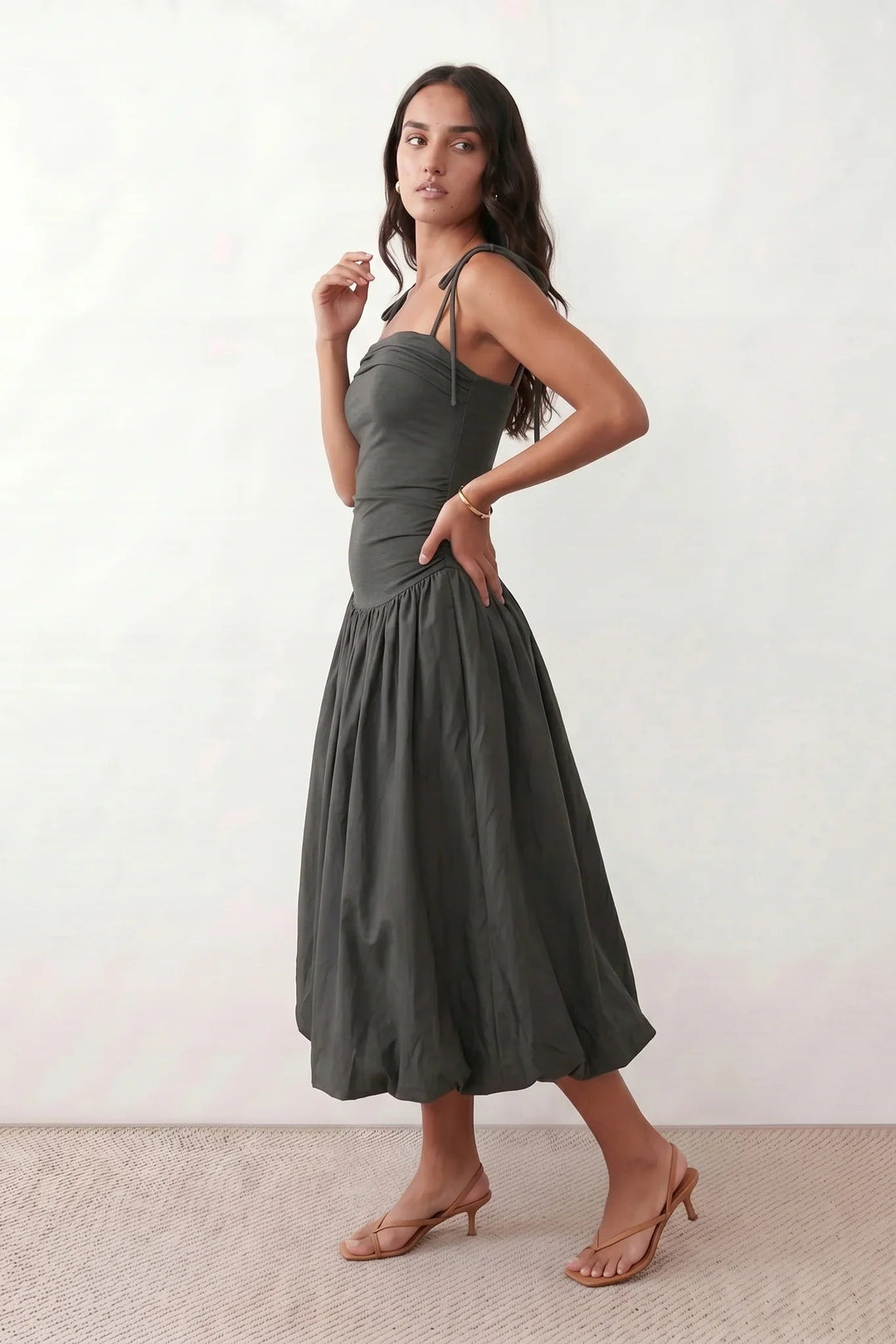 Woman wearing a spaghetti strap low back midi dress with a square neckline, ruched ruffle bodice, adjustable tie shoulder straps, elasticated smocked back, and flowing A-line skirt, styled as an elegant wedding guest and summer occasion dress.