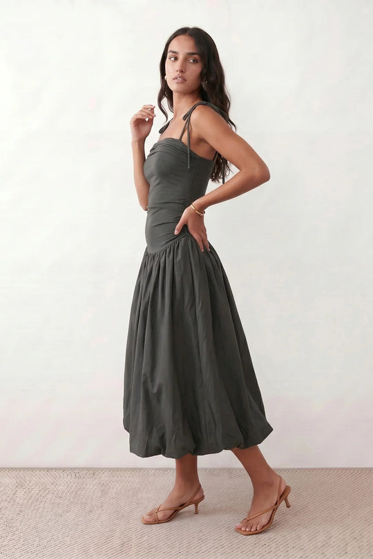 Woman wearing a spaghetti strap low back midi dress with a square neckline, ruched ruffle bodice, adjustable tie shoulder straps, elasticated smocked back, and flowing A-line skirt, styled as an elegant wedding guest and summer occasion dress.