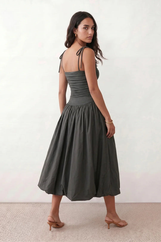 Woman wearing a spaghetti strap low back midi dress with a square neckline, ruched ruffle bodice, adjustable tie shoulder straps, elasticated smocked back, and flowing A-line skirt, styled as an elegant wedding guest and summer occasion dress.